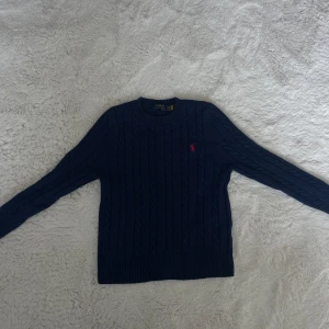 Mörkblå stickad tröja från Polo Ralph Lauren - Mörkblå stickad tröja från Polo Ralph Lauren med klassiskt kabelstickat mönster och röd broderad logga på bröstet. Rund halsringning och långa ärmar. Perfekt för en stilren och tidlös look.