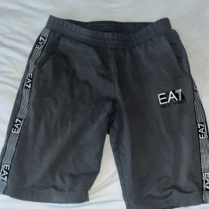 Svarta shorts från EA7 Emporio Armani - Snygga svarta shorts från EA7 Emporio Armani med vita ränder och logga längs sidorna. Shortsen har snörning i midjan och två sidofickor. Perfekta för en sportig och avslappnad stil.