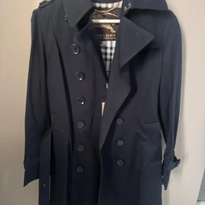 Mörkblå trenchcoat  - Säljer en klassisk mörkblå trenchcoat  med det ikoniska rutiga fodret. Jackan har dubbelknäppning, bälte i midjan och vid ärmslut samt stora slag. Perfekt för dig som vill ha en tidlös och stilren jacka.