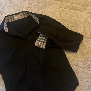 Svart skjorta med rutiga detaljer från Burberry - Stilren svart skjorta från Burberry med klassiska rutiga detaljer på insidan av kragen och ärmsluten. Skjortan har långa ärmar och knäppning framtill. Perfekt för dig som gillar en tidlös och elegant look.