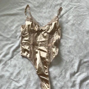 Champagnefärgad korsett från PrettyLittleThing - Snygg champagnefärgad korsett från PrettyLittleThing i glansig satin med spetsdetaljer och reglerbara axelband. Kuporna är formade och ger extra stöd. Perfekt för dig som vill ha en trendig och feminin look med lyxig känsla.