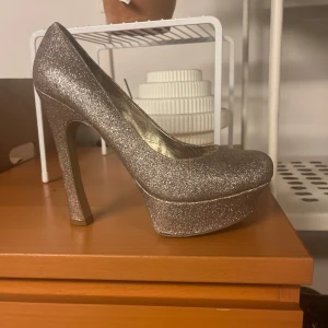 Glittriga silverfärgade pumps med platå - Säljer ett par snygga pumps i silver med glittrig yta och hög klack. Skorna har rund tå och en rejäl platåsula som ger extra höjd. Perfekta för dig som vill sticka ut på festen!