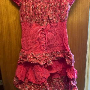 Rosa blommig kortklänning med volang och snörning - Supersöt rosa kortklänning med blommigt mönster och volangdetaljer. Klänningen har korta ärmar, smockad midja och snörning framtill. Perfekt för sommarens alla tillfällen! (Barn)