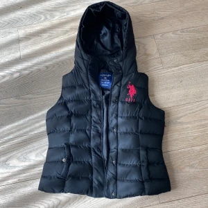 Svart dunväst med huva från U.S. Polo Assn. - Svart dunväst från U.S. Polo Assn. med huva och broderad röd logga på bröstet. Västen har tryckknappar och dragkedja framtill samt fickor på sidorna. Tillverkad i 100% polyester och har en sportig, klassisk look. I stort sett oanvänd.