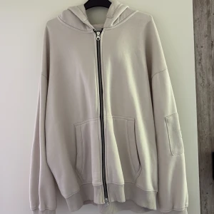 Beige hoodie med dragkedja från Bershka - Säljer en beige hoodie från Bershka i loose fit-modell. Tröjan har dragkedja framtill, huva och stora fickor. Detalj med patch på ena ärmen där det står 'Modern Archives'. Perfekt för en avslappnad stil.