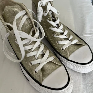 Glittriga guld Converse All Star - Snygga Converse All Star high-tops i glittrigt guld med vita snören och klassisk vit tå. Skorna har rund tå, platt sula och den ikoniska Converse-loggan på sidan. Perfekta för dig som vill sticka ut med lite extra bling i vardagen.