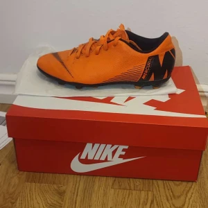 Nike Mercurial orange fotbollsskor - Snygga Nike Mercurial fotbollsskor i en riktigt poppig orange färg med svarta detaljer och snörning. Skorna har låg profil, platt sula med dobbar och är tillverkade i syntetmaterial för bästa grepp och känsla på planen. Perfekta för dig som vill sticka ut på fotbollsplanen.