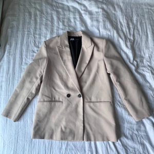 Beige kavaj från Zara - Stilren beige kavaj från Zara med klassisk krage och två knappar framtill. Kavajen har raka fickor och är helfodrad med svart innerfoder. Perfekt för en clean och modern look.