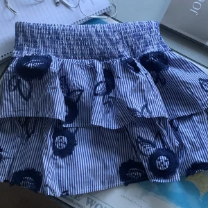 Randig volangkjol Holly & Whyte S - Superfin volangkjol från Holly & Whyte med blåvita ränder och broderade blommor i mörkblått 💙 Kjolen har bred smock i midjan och dubbla volanger för extra volym. Midjehöjden kan justeras 💓