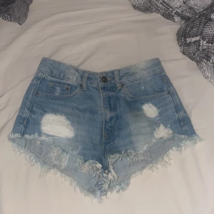 Blå jeansshorts med fransar från H&M Divided - Säljer ett par blå jeansshorts från H&M Divided i storlek 34. Shortsen har fransiga kanter och slitna detaljer framtill för en cool, somrig look. Klassisk femficksmodell med knapp och dragkedja.