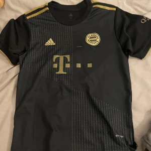 FC Bayern München svart fotbollströja med guldtryck - Snygg svart FC Bayern München fotbollströja från Adidas med guldiga detaljer och tryck. Tröjan har klubbmärke och sponsorlogga på framsidan samt spelarnamn och nummer 10 (Sané) på ryggen. Klassisk passform och korta ärmar.