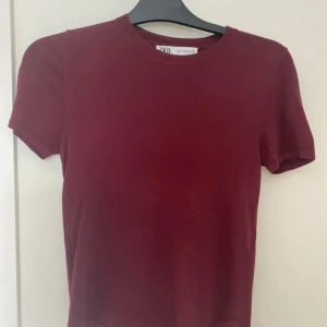 Vinröd topp från Zara i bomull - En vinröd, tight topp från Zara med rund halsringning och korta ärmar. Tillverkad i mjuk bomull som sitter snyggt mot kroppen. Perfekt att styla med jeans eller kjol för en clean look.
