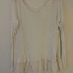 Vit volangtröja från MQ, 365 - Säljer en söt vit långärmad tröja från MQ,  365 med volangdetalj nedtill och spetskant. Tröjan har en lätt v-ringning och är gjord i ett tunt, mjukt material som känns luftigt och skönt.