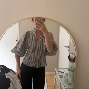 Randig skjorta med vida ärmar - Säljer en snygg skjorta med svarta och vita ränder och vida trekvartsärmar. Skjortan har knappar framtill och en avslappnad passform. Perfekt för dig som vill ha något stilrent men ändå unikt.