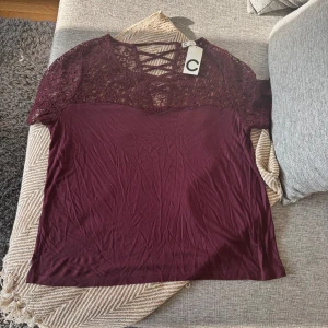 Vinröd spetsblus med snörning, Cubus  - Supersnygg vinröd blus från Cubus  med spets upptill och trendig snörning vid halsen. Blusen har korta ärmar och en mjuk, stretchig känsla i tyget. Perfekt för dig som gillar detaljer och vill sticka ut lite extra.