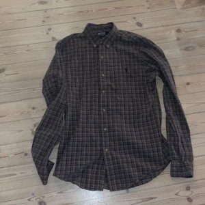 Rutig skjorta Polo Ralph Lauren M - Snygg rutig skjorta från Polo Ralph Lauren i slim fit-modell. Skjortan har lila, gröna och blå toner, klassisk krage och knäppning framtill. Tillverkad i mjuk bomull och har långa ärmar. Perfekt för dig som gillar stilrena och tidlösa plagg. Made in fillipines.