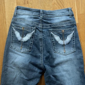 Blå bootcut jeans med broderade bakfickor - Säljer ett par blå bootcut jeans med snygga slitningar och broderade detaljer på bakfickorna. Jeansen har normal passform, dragkedja och knapp i gylfen. Perfekta för dig som gillar en klassisk men ändå unik look.