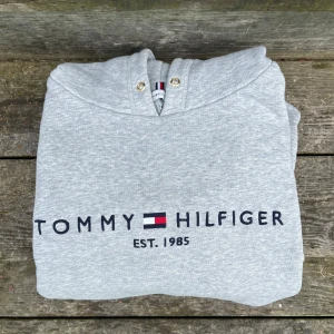 Grå hoodie från Tommy Hilfiger - Säljer en ljusgrå hoodie från Tommy Hilfiger med logga och texten 'EST. 1985' framtill. Tröjan har huva med guldfärgade öljetter och är tillverkad i mjukt bomullsmaterial. Perfekt för en avslappnad stil. Har tyvärr blivit för stor för den, den ligger bara och skräpar i garderoben😅