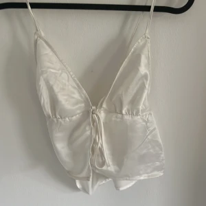 Vit satin linnetopp med knyt - Säljer en vit linnetopp i glansigt satinmaterial med smala axelband och djup v-ringning. Toppen har knyt framtill och är perfekt för sommarens varma dagar. Enkel och stilren design som passar till mycket.