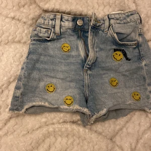 Jeansshorts med smiley-motiv från H&M x Smiley World - Ljusa jeansshorts med råa kanter och flera gula smiley-ansikten framtill. Ena bakfickan är gul med ett stort svart smiley-tryck. Klassisk femficksmodell och bälteshällor. Perfekta för en lekfull stil.
