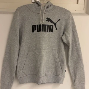 Grå hoodie från Puma - Säljer en klassisk grå hoodie från Puma med svart logga på bröstet. Tröjan har huva med dragsko och en stor magficka framtill. Perfekt för en avslappnad stil och enkel att matcha med det mesta.