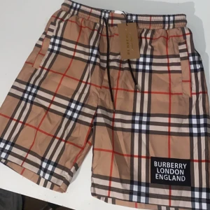 Rutigta Burberry shorts med snörning - Snygga shorts från Burberry i klassiskt beige rutigt mönster med svarta, vita och röda detaljer. Shortsen har snörning i midjan, sidofickor och en patch med 'Burberry London England' framtill. Perfekta för sommaren. Har haft på mig de 1 gång men de är helt nya.