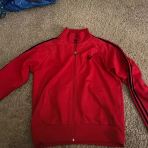Röd track jacket från Fred Perry - Säljer en klassisk röd track jacket från Fred Perry med svarta ränder längs ärmarna och broderad logga på bröstet. Jackan har dragkedja framtill, ståkrage och ribbade muddar. Perfekt för en sportig och stilren look. Ca Stl M