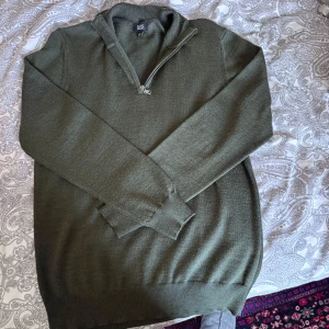 Olivgrön stickad tröja med dragkedja från Long Island - Säljer en olivgrön stickad tröja från Long Island med halv dragkedja vid halsen. Tröjan har lång ärm och ribbade muddar vid ärmslut och nederkant. Perfekt för lager-på-lager under kyliga dagar.