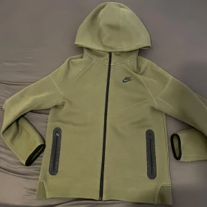 Grön Nike Tech flecce - Nike med svart logga på bröstet. Jackan har hel dragkedja framtill, två fickor med dragkedja och huva. Perfekt för en sportig och avslappnad stil.