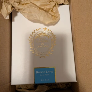 Bianco Latte Eau de Parfum från Giardini di Toscana - Exklusiv parfym i elegant vit kartong med gulddetaljer och blå etikett. Bianco Latte är en Eau de Parfum på 100 ml, tillverkad i Italien. Förpackningen har en lyxig känsla med klassisk design och tydlig märkning. Diskuterar inte priset.