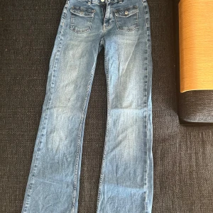 Ljusblå jeans från Kappahl, stl 164 - Snygga ljusblå jeans från Kappahl i klassisk utsvängd modell. Jeansen har två framfickor med lock och knapp, samt två vanliga bakfickor. Tillverkade i mjukt denim med dragkedja och knapp framtill. Perfekta för en avslappnad och trendig look. 