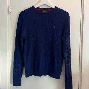 Blå kabelstickad tröja från Morris - En helt ny blå kabelstickad tröja från Morris i 100% merinoull. Strl S. Tröjan har rund halsringning, lång ärm och en liten röd morris logga på bröstet. Perfekt för dig som gillar klassisk och stilren outfit.