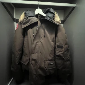 Brun dunjacka från Canada Goose  - Säljer en brun Canada Goose dunjacka med fet pälskant på huvan och klassisk röd logga på ärmen. Jackan har stora fickor, dragkedja och snörning i huvan. Perfekt för kalla vinterdagar. Stilen är loose och ärmarna är långa. Har slitage på armarna men det går enkelt att fixa.