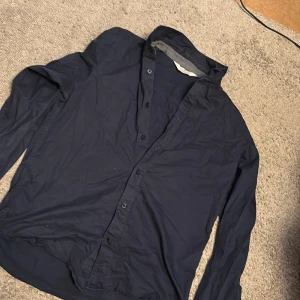 Mörkblå skjorta från H&M - Snygg mörkblå skjorta från H&M med klassisk krage och knäppning framtill. Skjortan har långa ärmar och är tillverkad i ett lätt och bekvämt material, perfekt för en stilren look. Passar dig som gillar enkel och tidlös stil.