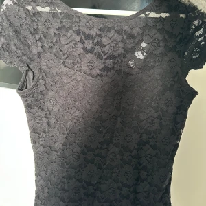 Svart spets blus från Vila - Säljer en svart klänning från Vila i storlek XS. Blusen har korta ärmar och är helt täckt av ett vackert blommigt spetsmönster. Perfekt för dig som gillar en stilren look. I mycket bra skick därav använd endast  1 gång , säljer då den bara ligger i garderoben🥰❤️‍🔥 skicka vid fler frågor och mm. Annars är det bara att köpa! Första bilden är framsidan och andra bilden är baksidan ❤️ser ut som en klänning på bilderna men OBs det är en Blus 👚 