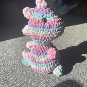 regnbågig dinosaurie  - Det här är en virkad, färgglad dinosaurie i pastellfärger. Den är mjuk och har detaljer som taggar på ryggen och svansen. Perfekt som gosedjur eller dekoration för unga och unga vuxna som gillar söta och unika handgjorda saker.