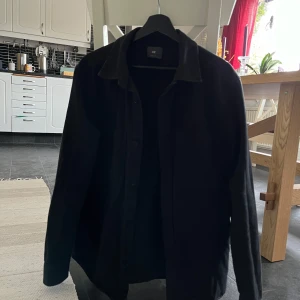 Svart overshirt från H&M - Säljer en svart overshirt från H&M med klassisk krage och bröstficka. Skjortan har långa ärmar och knäppning framtill. Perfekt att bära som lager över en t-shirt eller tröja.
