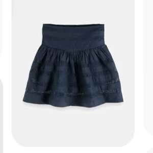 Marinblå kortkjol med volangdetaljer - Svart kortkjol med bred, smockad midja och volangiga partier med shorts under. Kjolen har dekorativa spets- och sömdetaljer som ger en bohemisk känsla. Perfekt att styla med en enkel topp eller hoodie.köpte för 700kr