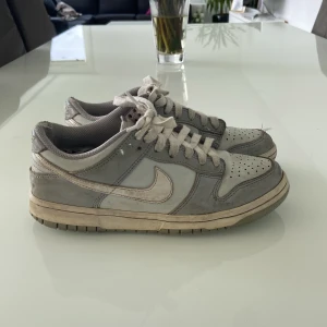 Nike Dunk sneakers i grått och vitt - Nike Dunk sneakers i grått och vitt med klassisk låg profil. Skorna har snörning, rund tå och platt sula. Perfekta för dig som gillar en stilren och sportig look. Swoosh-logga på sidorna och Nike-brodyr på hälen.