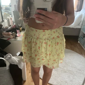 Gul blommig volangkjol - Supersöt gul kjol med blommigt mönster och volanger. Kjolen har smock i midjan för en bekväm passform och är kort i modellen. Perfekt för varma dagar och ger en romantisk vibe till outfiten.