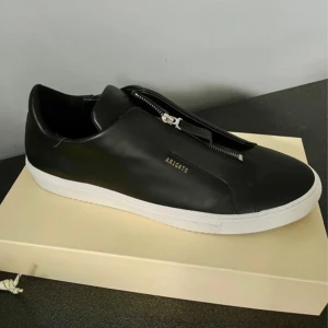 Svarta sneakers med dragkedja från Axel Arigato - Stilrena svarta sneakers från Axel Arigato med vit sula och dragkedja på ovansidan. Skorna har minimalistisk design med diskret logga i guld på sidan. Perfekta för dig som gillar en clean look. Skinn 100%. Använda 2 gånger. Nypris 2900. Passar 45 och 46. 