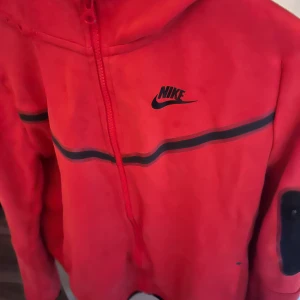 Röd Nike zip-hoodie med svarta detaljer - Röd hoodie från Nike med dragkedja och svarta ränder över bröstet och ärmarna. Klassisk Nike-logga på vänster sida. Huva och fickor framtill, samt svarta detaljer på ärmarna.