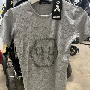 Grå t-shirt med mönster och nitar från Philipp Plein - Snygg grå t-shirt från Philipp Plein med ett diskret spetsliknande mönster och dekorativa nitar som formar märkets logotyp på bröstet. Rund halsringning och korta ärmar. Perfekt för dig som gillar statement-plagg.