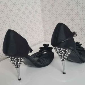 Svarta pumps med strass och blomdetalj från Fusion - Säljer ett par svarta eleganta pumps från Fusion med öppen tå, smal klack och strassdetaljer på hälen. Skorna har en stor tygblomma framtill och ett elegant band runt vristen. Perfekta för dig som vill sticka ut lite extra.