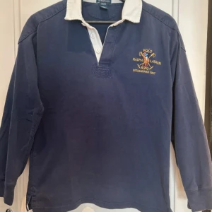 Ralph lauren rugby tröja  - Säljer en klassisk blå rugbytröja från Polo Ralph Lauren med vit krage och broderad logga på bröstet. Tröjan har långa ärmar och en stor röd siffra 8 på ryggen. Tillverkad i 100% bomull och passar perfekt för en sportig stil. Storlek L barn vilket bör passa som XS längden på tröjan är 63cm