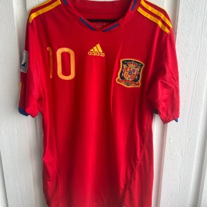 Spansk landslagströja Fabregas 10 Adidas - Röd fotbollströja från Spaniens landslag med gula detaljer, Adidas-logga och nummer 10. Fabregas namn och nummer på ryggen. Korta ärmar och broderat landslagsmärke på bröstet. Perfekt för fotbollsfans!