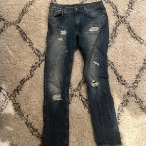 Blå slitna jeans från Nudie Jeans - Säljer ett par blå jeans från Nudie Jeans med flera slitningar och lagade detaljer framtill. Klassisk femficksmodell med raka ben och normal passform. Perfekta för dig som gillar en lite ruffare stil. Skick 9/10