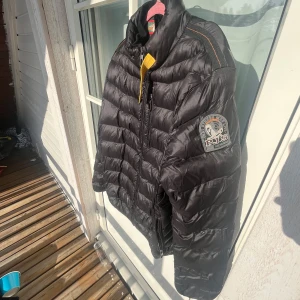 Svart dunjacka med patch på ärmen - Säljer en svart dunjacka från parajumpers med markerade sömmar och patch på ena ärmen. Jackan har dragkedja framtill och hög krage. Perfekt för kyligare dagar och lätt att bära tack vare sin lätta konstruktion.
