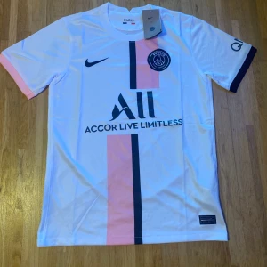 PSG vit fotbollströja med rosa och svart detalj - Säljer en vit PSG fotbollströja från Nike med rosa och svart vertikal rand på framsidan. Tröjan har korta ärmar, PSG-logga på bröstet och sponsortryck. Perfekt för dig som älskar fotboll och vill visa ditt stöd för Paris Saint-Germain.
