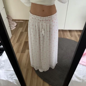 Vit långkjol med blommigt mönster från Gina Tricot - Supersöt vit långkjol från Gina Tricot med små rosa blommor och smock i midjan. Kjolen har knytband med tofsar och ett luftigt, flowy tyg som passar perfekt till varmare dagar. Använd få gånger!! Säljer för 90kr. Säljer eftersom att den är för kort